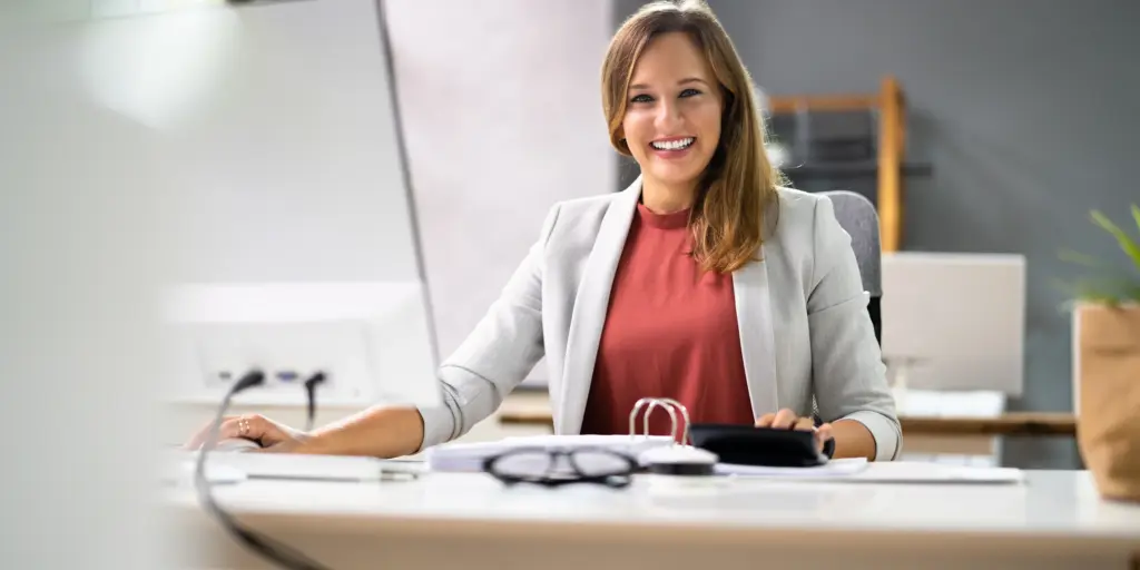 Eine lächelnde Frau im Büro am Schreibtisch vor einem Laptop sitzend, trägt ein rotes Oberteil und einen grauen Blazer.