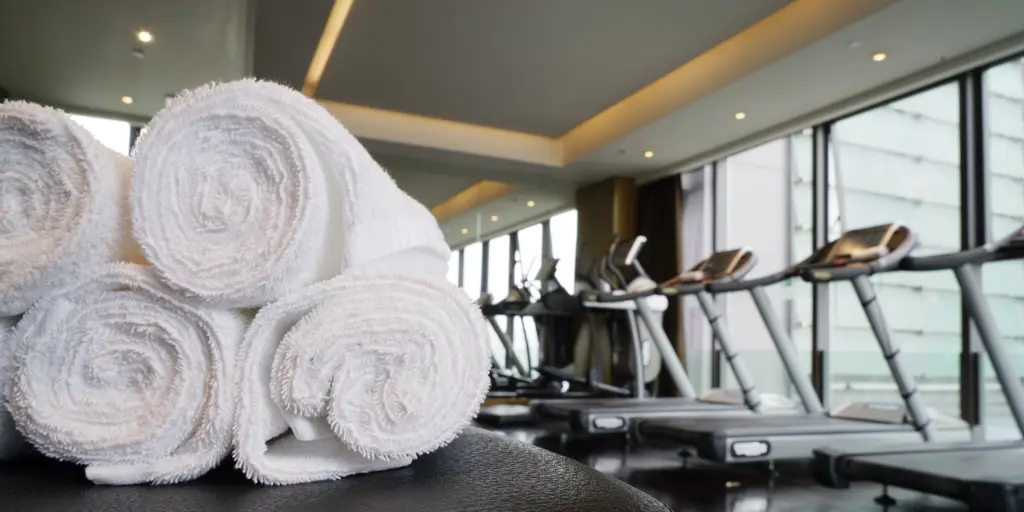 Weiße Handtücher auf einer Bank im Fitnessstudio mit Laufbändern im Hintergrund.