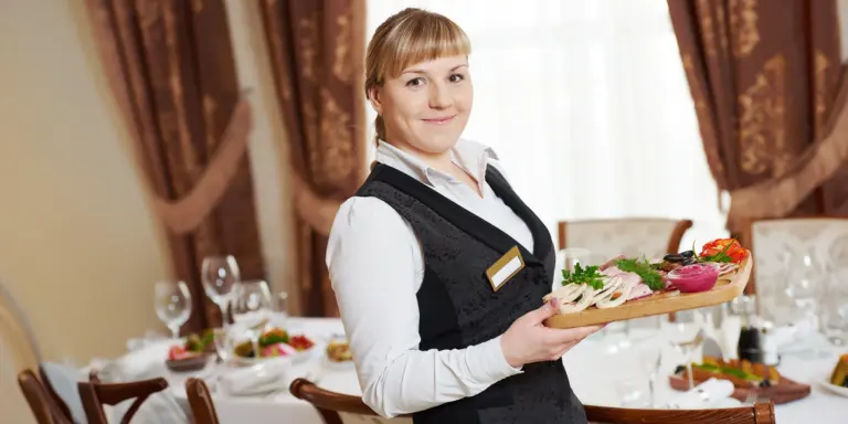 Kellnerin mit Servierplatte im eleganten Restaurant, gedeckter Tisch im Hintergrund.